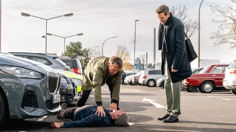 Achim Weishaupt (Torsten Knippertz, liegend) verteilt Flyer für einen kölschen Verein auf dem Parkplatz des SOKO-Präsidiums. Als Kommissare Matti Wagner (Pierre Besson, l.) und Jonas Fischer (Lukas Piloty, r.) eintreffen, bricht er plötzlich vor ihnen zusammen – und stirbt. – Bild: ZDF und Martin Rottenkolber