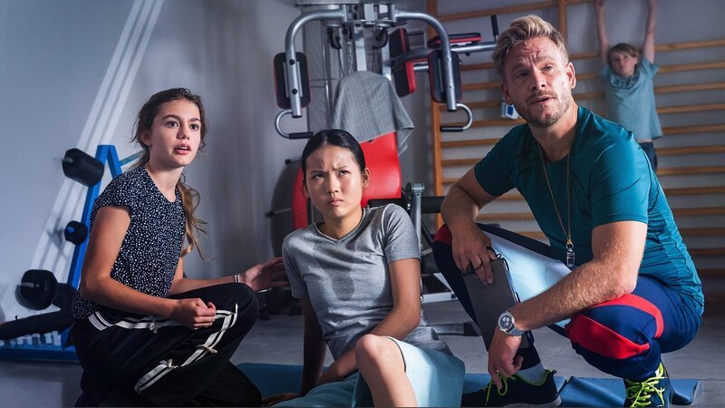 Suki (Alva Müllenberg, links), Anh (Ha My Anna Tran, Mitte) und Sportlehrer Hauser (Frederic Heidorn, rechts) schauen genervt zu Joshua. – Bild: MDR/​Thomas Dietze /​ KiKA