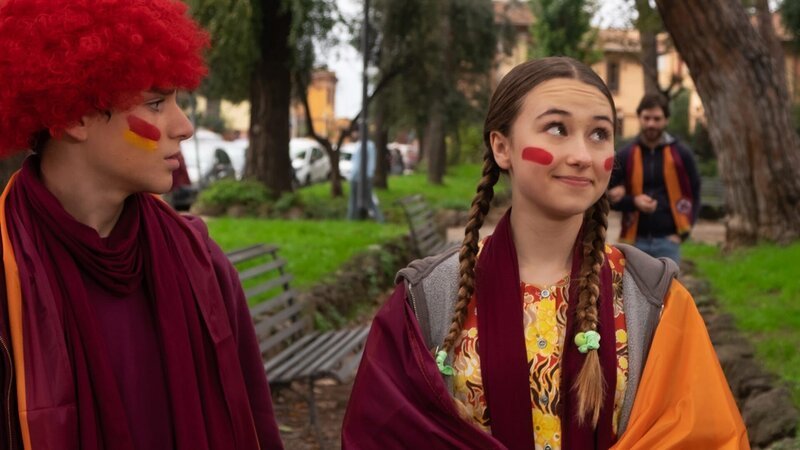 Lucy (Kensington Tallman, r.) und Nico (Christian Monaldi, l.) tragen Schals und Perücken in den Farben ihrer Fußballmannschaft – orange und rot. – Bild: hr/​Red Monk Studios srl/​ HSR S1 (FGM) Inc.