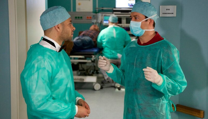 Dr. Philipp Brentano (Thomas Koch, rechts) untersucht gerade einen Patienten (Veit Stübner, hinten links) mit einem abgetrennten Daumen, als plötzlich Dr. Matteo Moreau (Mike Adler, links) im Frack auftaucht und seine Hilfe anbietet. Da Moreau sehr präsent im Behandlungszimmer erscheint, bittet Brentano ihn kurz vor die Tür. Dr. Matteo Moreau ist „der“ Handspezialist aus dem Johannes-Thal-Klinikum Erfurt und versucht sich um den Ärzteball zu drücken. Er fragt Brentano höflich, ob er ihn bei der Behandlung unterstützen könne und vergisst nicht zu erwähnen, dass er das Beste ist, was Brentano jetzt gerade passieren könne. – Bild: MDR/​Rudolf Wernicke
