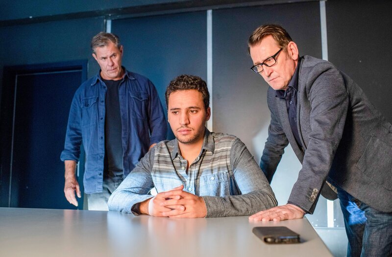 Der junge Jassim Abdaly (Mo Issa, M.) sitzt im Vernehmungsraum der Kripo Lübeck. Die beiden Ermittler Finn Kiesewetter (Sven Martinek, l.) und Lars Englen (Ingo Naujoks, r.) versuchen ihn zum Reden zu bringen. – Bild: ORF/​ONE GATE/​ARD/​Thorsten Jander
