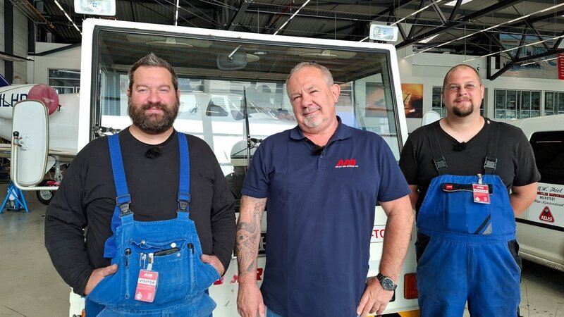 Marcus Thiel, his colleague Florian „Blitzi“ Hörig, and Tool Shop Manager Mike Streif pose next to an aircraft tractor in the workshop hall of the Atlas Air Service aircraft hangar in Bremen. – Bild: Warner Bros. Discovery, Inc. or its subsidiaries and affiliates. All Rights Reserved. /​ Berit Hinnerks