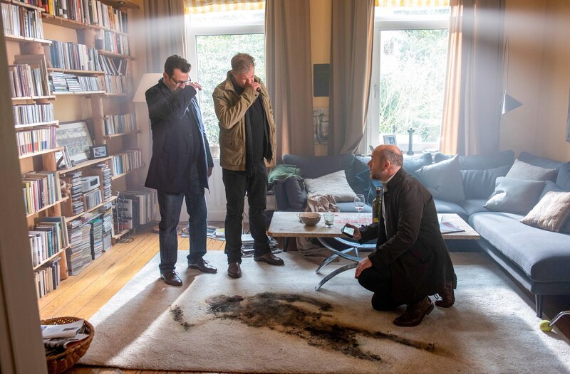 Die Kommissare Finn (Sven Martinek, l.) und Lars (Ingo Naujoks, 2.v.l.) stehen am Fundort der Leiche von Hans Vonnsen. Auf dem Teppich vor ihnen ist ein dunkler Abdruck des bereits stark verwesten Körpers zu sehen. Dr. Henning Strahl (Christoph Tomanek, r.) kniet auf dem Boden und dokumentiert den Tatort mit seiner Kamera. – Bild: ORF/​ONE GATE/​ARD/​Thorsten Jander