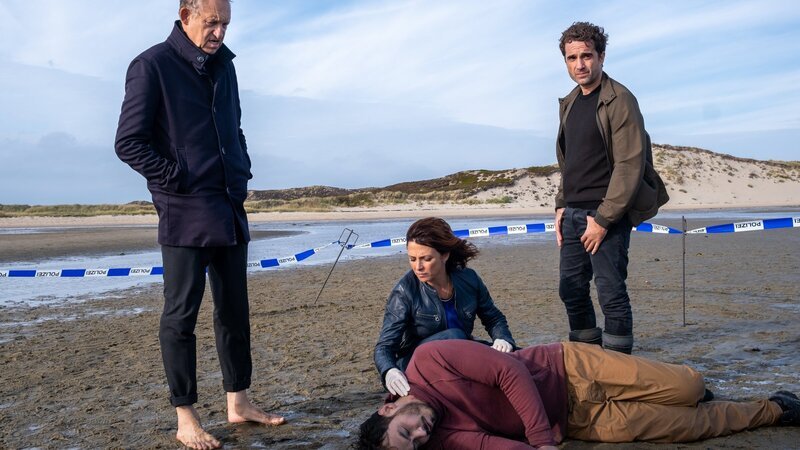 Die Kommissare Carl Sievers (Peter Heinrich Brix, l.), Ina Behrendsen (Julia Brendler) und Hinnerk Feldmann (Oliver Wnuk, r.) begutachten die Leiche von Pablo Hernandez (Iskander Madjitov, vorne liegend). Der Tote wurde mitten im Watt gefunden. – Bild: ZDF and Manju Sawhney