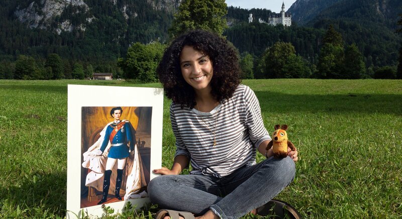 Moderatorin Siham El Maimouni mit einem Bild von König Ludwig vor dem Schloss Neuschwanstein – Bild: WDR/​gomie