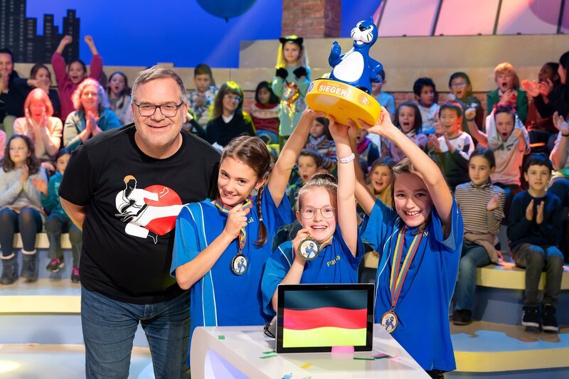 Das blaue Rateteam aus Waldbrunn/​Deutschland hat Grund zum Jubeln: Es kann siegreich den begehrten Piet-Flosse-Pokal in die Luft strecken! – Bild: ZDF und Ralf Wilschewski