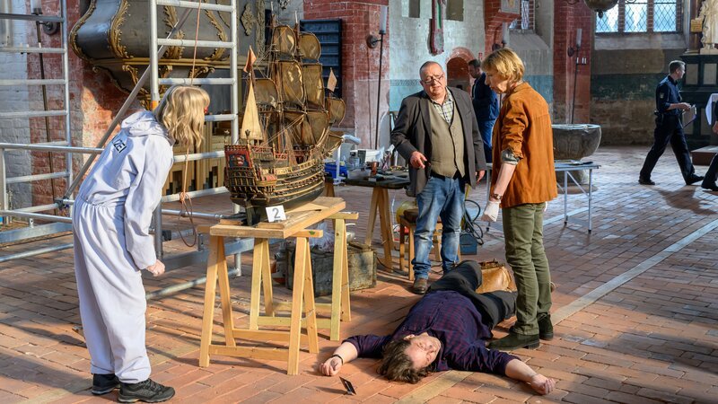 Erste Untersuchungen der Rechtsmedizinerin Dr. Helene Sturbeck (Katharina Blaschke, r.) ergeben, dass Sven Lundquist (Øystein Kloster, 2.v.r.) erschlagen wurde, gleich neben dem Modellschiff „Prerow“. In diesem findet Roswitha Prinzler (Silke Matthias, l.) Konfetti. Jan-Hinrich Reuter (Udo Kroschwald, 3.v.r.) hört den Ausführungen seiner Kolleginnen zu. – Bild: ZDF/​Meyerbroeker