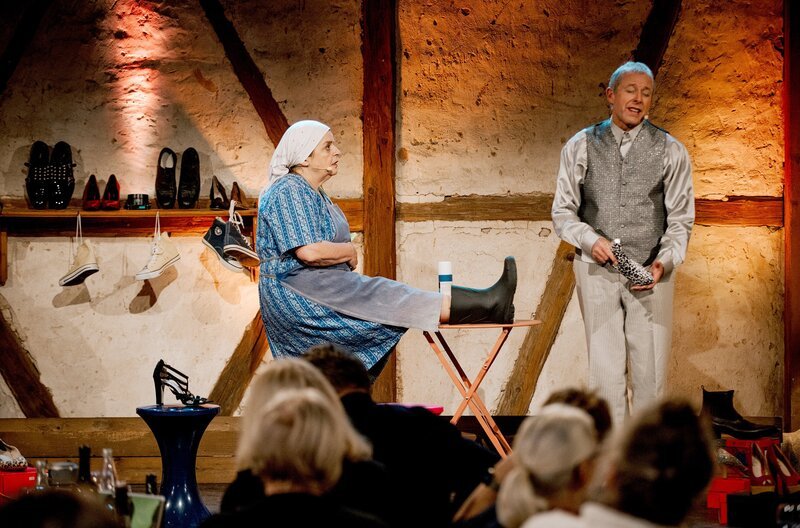 Bevor sich Bäuerin Lena (Hillu’s Herzdropfa) bei dem Verkäufer (Franz Auber /​ Hillu’s Herzdropfa) neue Schuhe kauft, bleibt sie lieber bei ihren treuen Gummistiefeln. – Bild: SWR/​Ronny Zimmermann
