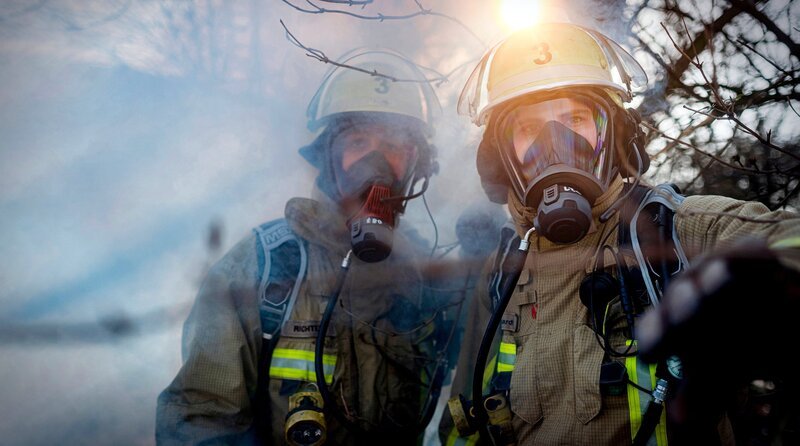Zwei Einsatzkräfte der Feuerwehr Duisburg mit Atemschutzmasken auf einem verrauchten Gelände. Eine schwarze Rauchwolke steigt über einem verlassenen Vereinsgelände auf, als der Löschzug der Wache 3 eintrifft. – Bild: WDR /​ Sebastian Drolshagen