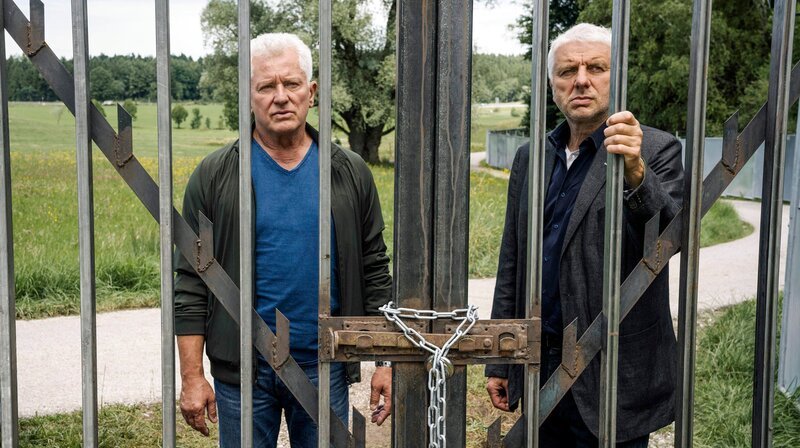 Von links: Die Kriminalhauptkommissare Ivo Batic (Miroslav Nemec) und Franz Leitmayr (Udo Wachtveitl) stehen vor dem verschlossenen Eingangstor zum „Freiland“-Gelände. – Bild: BR/​Hendrik Heiden/​Claussen+Putz Filmproduktion GmbH