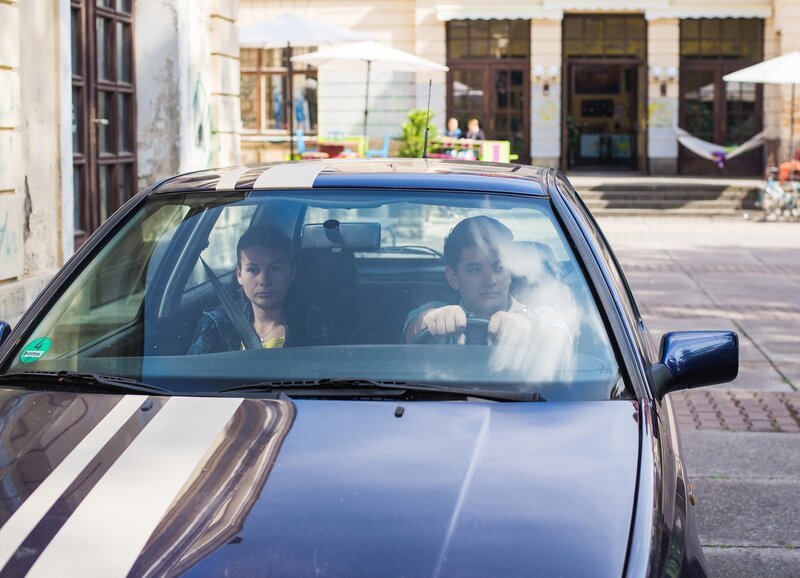 Orkan (Flavius Budean) hat sich ein Auto gekauft und fährt mit Jule (Maja Hieke) auf dem Internatshof vor. – Bild: MDR/​Saxonia Media/​Christoph Gorke
