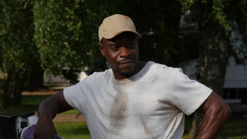 Joe leaning against a truck wearing a work shirt, gloves and holding work gloves. – Bild: TLC