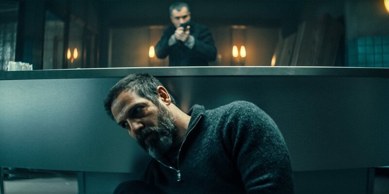 Reza (Yousef Sweid) versteckt sich vor dem Auftragskiller Brynner (Julian Sharp) hinter dem Vorbereitungstisch in der Bestattung. – Bild: ORF/​Mona Film/​ Barry Films