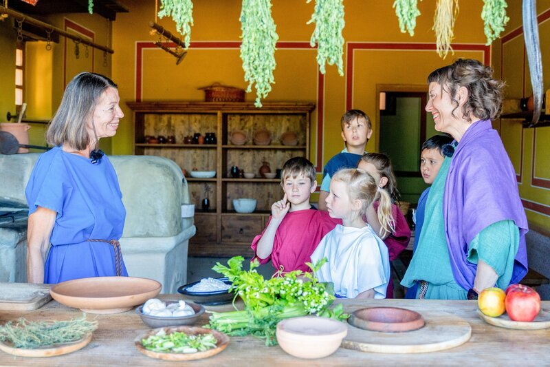 Köchin Antonia, die im echten Leben Anneliese heißt, zeigt den Kindern und Esther wie im Alten Rom Brot gebacken wurde. – Bild: ORF/​Römerstadt Carnuntum/​T. Mauerhofer
