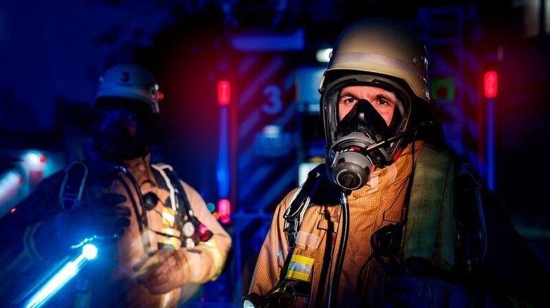 Zwei Einsatzkräfte der Feuerwehr Duisburg mit Atemschutzmasken im Dunkeln vor einem Einsatzfahrzeug. Die Duisburger Feuerwehr wird zu einem Gebäudebrand alarmiert: Ein Wohnungsbrand im Mehrfamilienhaus mit starker Rauchentwicklung im Treppenhaus. – Bild: WDR/​Sebastian Drolshagen