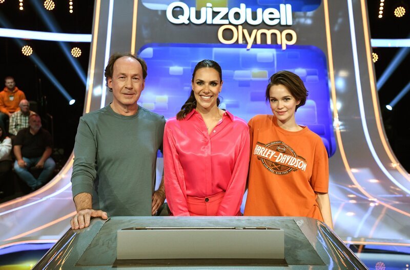 Quizmasterin Esther Sedlaczek (M.) mit den Kandidat:innen des Teams „Ku’damm“:  Ulrich Noethen (l.), Schauspieler und Emilia Schüle (r.), Schauspielerin. – Bild: ARD/​Uwe Ernst