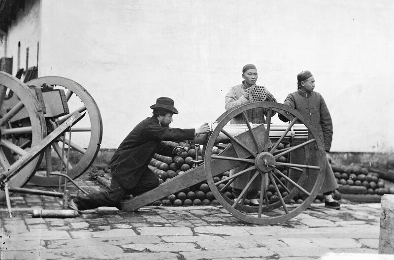 John Thomson (li.) posiert an einer Gatling-Kanone, bereit die Waffe abzufeuern. Neben ihm stehen zwei chinesische Männer, von denen einer den Verschluss der Waffe hält. – Bild: Wellcome Library, London