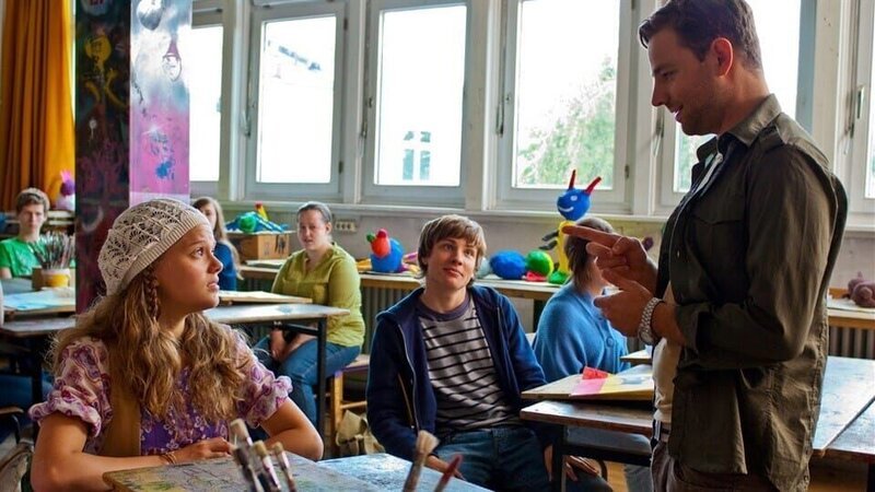 Nicolas (Jannik Schümann) amüsiert sich, weil Emma (Sophie Karbjinski) schon wieder wegen der fehlenden Kunstarbeit ermahnt wird. v.l.: Emma (Sophie Karbjinski), Nicholas (Jannik Schümann), Lehrer (Patrick Monkeberg). – Bild: NDR/​Southern Star