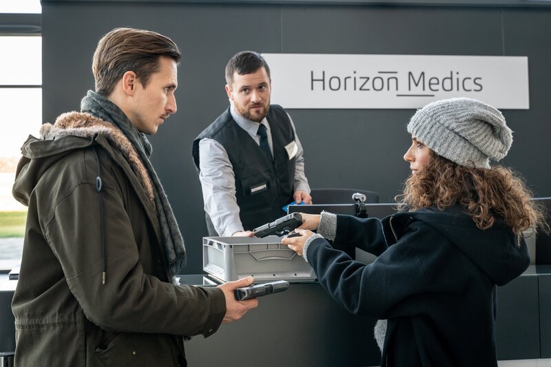Erste Ermittlungen führen zum ehemaligen Arbeitgeber der Toten: Horizon Medics. Kim Nowak (Amy Mußul, r.) und Moritz Brenner (Johannes Hendrik Langer, l.) sind über die Sicherheitsvorkehrungen des Unternehmens überrascht. Der Security-Mann Renè Makowski (Lorris André Blaszejewski, M.) bittet die beiden ihre Dienstwaffen abzugeben. – Bild: Uwe Frauendorf /​ ZDF