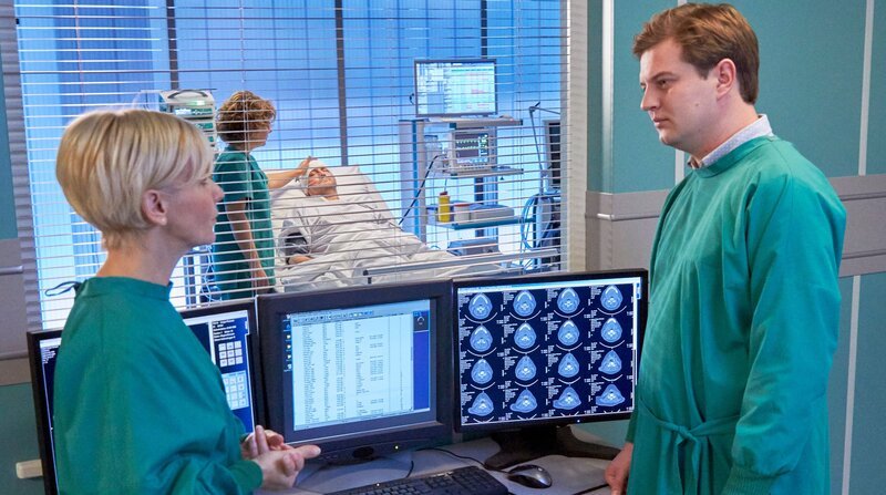 Jakob Heilmann (Karsten Kühn, re.) hat sich in der Cafeteria in den Finger geschnitten. Dr. Kathrin Globisch (Andrea Kathrin Loewig, li.) hat die kleine Wunde schnell versorgt. Dabei sehen die beiden einen Mann (Ingo Naujoks, hi.re.), der ohne Papiere nach einem Unfall eingeliefert wurde und um den sich Schwester Ulrike (Anita Vulesica, hi. li.) kümmert. Jakob kommt der Mann irgendwie bekannt vor, doch er kann sich nicht erinnern, woher. – Bild: MDR/​Saxonia Media/​Kiss