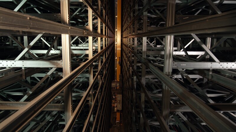Das Bild zeigt das komplexe Innere eines vollautomatisierten Hochregallagers, das als Aushängeschild der modernen intelligenten Logistik gilt. Diese spezielle Ansicht verdeutlicht die hochverdichteten Lagerregale und den zentralen Gang, in dem autonome Roboter oder automatische Lager- und Bereitstellungssysteme (AS/​RS) im Einsatz sind. – Bild: 14a Media GmbH