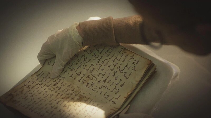 Eine behandschuhte Hand hält ein altes Notizbuch mit dicht beschriebenen Seiten. – Bild: The Movie Database (TMDb)