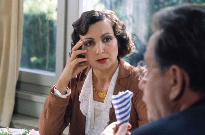 Im Mordfall „Marzipankartoffel“ wird die lebenslustige Putzmacherin Apollonia Schmaus (Ewa Teilmans) von Amtsmann Grüner (Gerd Fritz) verhört. – Bild: BR/​Tellux-Film GmbH