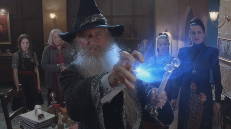 Der Große Magier (Nicholas Jones) ist an die Akademie gekommen, um das Rätsel um die gefälschten Unterschriften zu lösen. Mildred (Bella Ramsey, l.), Frau Graustein (Clare Higgins, 2.v.l.), Esther (Jenny Richardson, 2.v.r.) und Frau Harschmann (Raquel Cassidy, r.) sind gespannt, was der Zauber ihnen offenbaren wird. – Bild: ZDF und BBC/​ZDF Enterprises