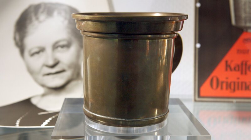 Der erste Melitta-Kaffeefilter und ein Foto seiner Erfinderin Melitta Bentz in einer Vitrine des Melitta-Archivs. – Bild: WDR/​dpa
