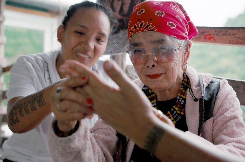 Während Whang Od aus der Hand einer Tattoo-Kundin liest, schaut ihre Großnichte Grace aufmerksam zu und lernt. Sie soll die Nachfolge der Matriarchin antreten und die Batok-Tradition in die Zukunft tragen. – Bild: André Krüger