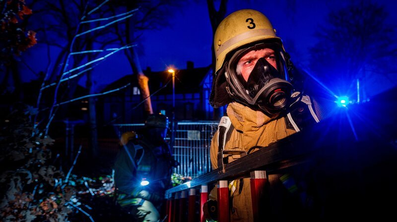 Zwei Feuerwehrmänner im Dunkeln mit Atemschutzmaske tragen eine Leiter. Rückblick Jahreswechsel: Für die Feuerwehr Duisburg traditionell eine der härtesten Nächte des Jahres. Die Einsatzkräfte stellen sich auf Dauerstress ein. – Bild: WDR/​Sebastian Drolshagen