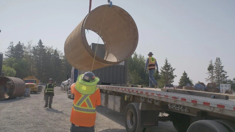 Die Arbeiter auf der Baustelle heben mithilfe eines Krans ein großes zylindrisches Betonelement von einem Lastkraftwagen an. – Bild: The Movie Database (TMDb)