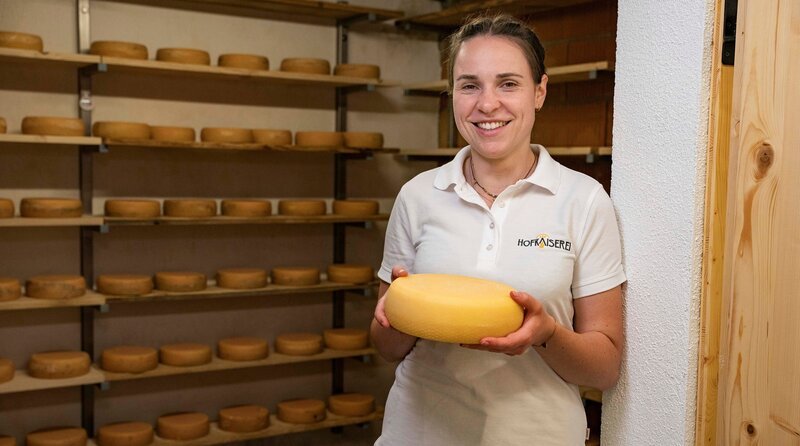Magdalena Kaiser steht in ihrem Reiferaum und hält einen Laib selbstgemachten Käse in der Hand. – Bild: BR/​megaherz gmbh/​Moritz Sonntag
