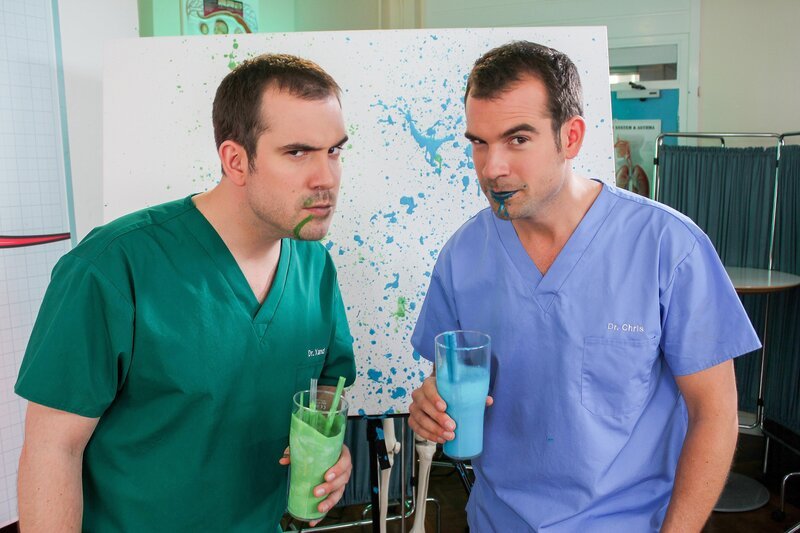 Dr. Xand (l., Xand van Tulleken) und Dr. Chris (r., Chris van Tulleken) vor einer Leinwand, auf der die gesprenkelten Flecken die Verbreitung von Viren beim Niesen zeigen. – Bild: ORF/​Serious Kids