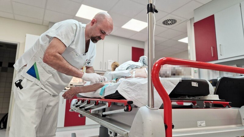 E6 – Male bald nurse placing an access inlay in the patient’s arm. The red handle bars are in the front of the pic, the patient is blurred – Bild: Warner Bros. Discovery, Inc.