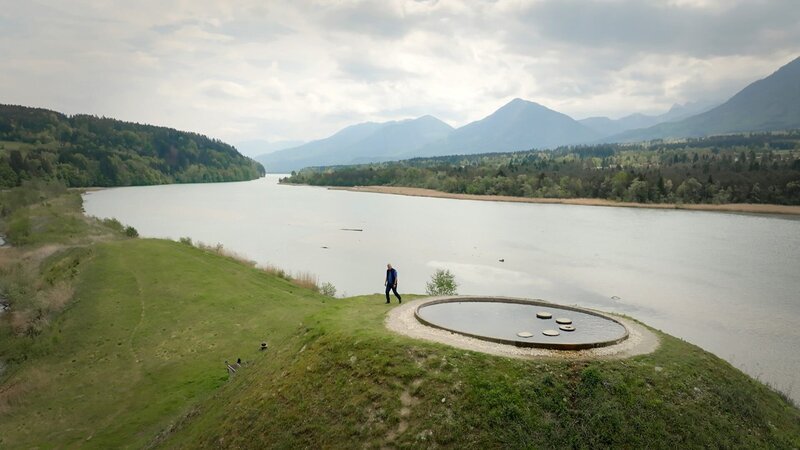 Eine Person bewegt sich durch eine Landschaft mit einem See im Tal, grünen Hängen und Bergen im Hintergrund, in der Nähe einer kreisförmigen Wasserinstallation – Bild: Wega Film