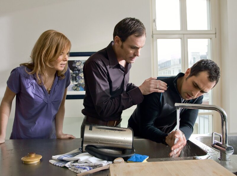 Ralf Meinecke (Gabriel Merz, rechts) hat sich bei seinem befreundeten Pärchen Valerie (Milena Dreißig, links) und Konrad Gerstenmeyer (Anian Zollner) mit kochend heißem Wasser die Hand verbrannt. – Bild: MDR/​Saxonia/​Peter Krajewsky