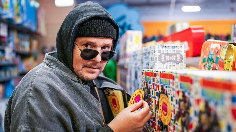Stefan Steindl (Thomas Mraz) klebt Plastik-Warnkleber auf Spielzeugverpackungen in einem Spielzeuggeschäft im Einkaufszentrum. Weiteres Bildmaterial finden Sie unter www.br-foto.de. – Bild: BR Stefan Steindl (Thomas Mraz) klebt Plastik-Warnkleber auf Spielzeugverpackungen in einem Spielzeuggeschäft im Einkaufszentrum. Weiteres Bildmaterial finden Sie unter www.br-foto.de. – Bild: BR
