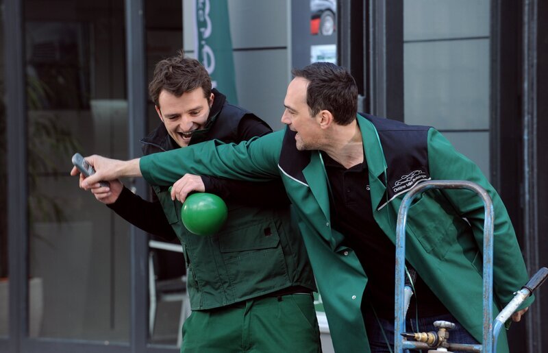 Mit vom Helium piepsiger Stimme beantwortet Flori das Werkstatttelefon. Von links: Florian Brunner (Tommy Schwimmer) und Mike Preissinger (Harry Blank). – Bild: BR/​Marco Orlando Pichler
