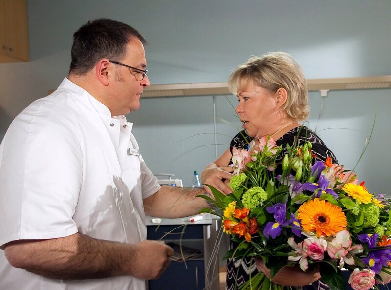 Pfleger Hans-Peter Brenner (Michael Trischan) bringt Sabine Schneider (Daniela Hoffmann) Blumen. Sabine Schneider ist äußerst angetan von Hans-Peter, dem das allerdings ziemlich unangenehm ist und er nicht weiß, wie er aus dieser Situation herauskommt. Doch plötzlich setzt bei Sabine Schneider wieder starkes Herzrasen ein – und das hat nichts mit Hans-Peter zu tun. – Bild: MDR/​MDR/​Krajewsky