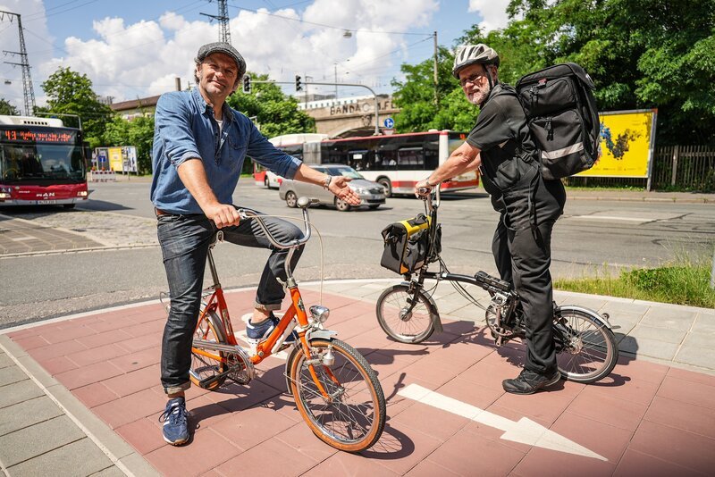 Der Schmidt Max (links) hat das alte Klapprad aus dem Keller geholt und begibt sich mit seinem orangen Oldtimer auf eine Reise durch Franken. Unterwegs trifft er Menschen auf ihren Falträdern; u.a. der Nürnberger Kaminkehrer Bernd Thiel (rechts). – Bild: BR/​André Goerschel