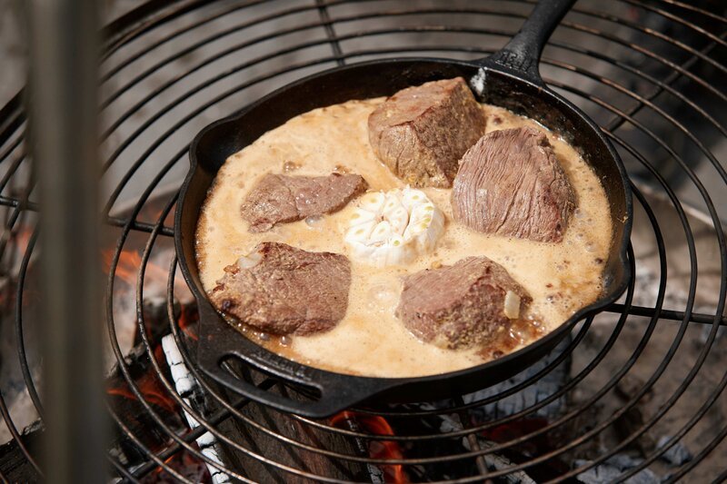 Bisonschulter in Biersoße mit geschmortem ganzen Knoblauch köchelt in der Pfanne auf dem Grill. – Bild: Marian Mok /​ BR, Yalla Productions GmbH /​ BR/​Yalla Productions GmbH/​Marian Mok