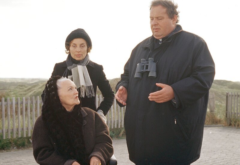 Pfarrer Braun (Ottfried Fischer) besucht die reiche Witwe Groenewold (Eva-Maria Hagen, im Rollstuhl) und ihre Pflegerin Maria (Teresa Harder). – Bild: ARD/​Degeto/​Romano Ruhnau