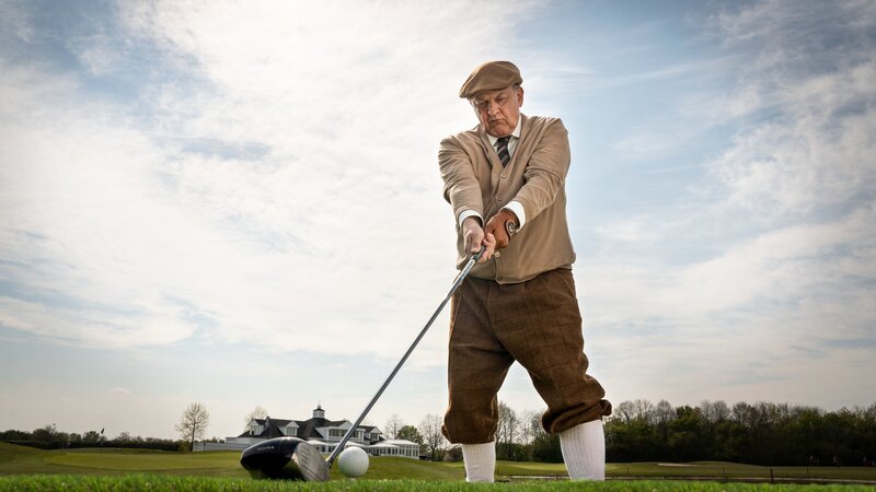 Privatdetektiv Georg Wilsberg (Leonard Lansink) beim Abschlag auf dem Golfplatz. Mit Schiebermütze, Knickerbockern und Strickjacke, spricht er den Ball an – fast so wie Gerd Fröbe in „James Bond – Goldfinger“. – Bild: ZDF/​Thomas Kost