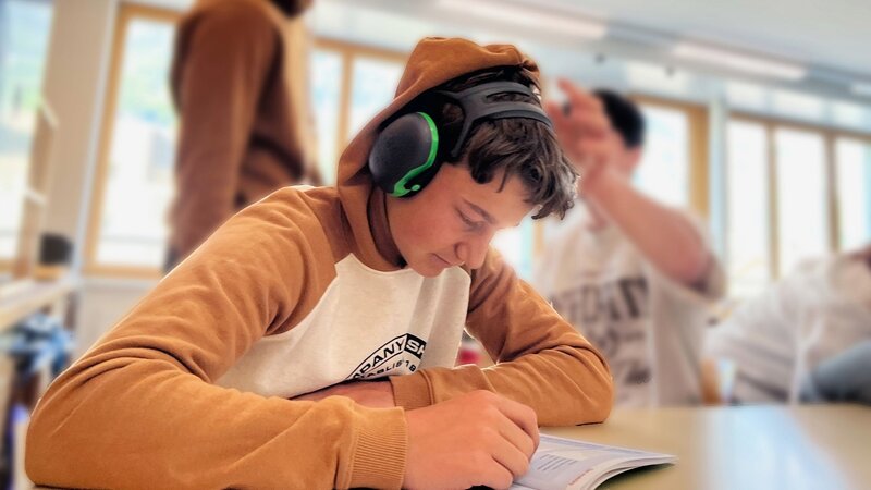 Ein Schüler sitzt mit Kopfhörer in der Klasse und versucht sich zu konzentrieren während der anderen Schüler ihn ablenken. – Bild: SRF