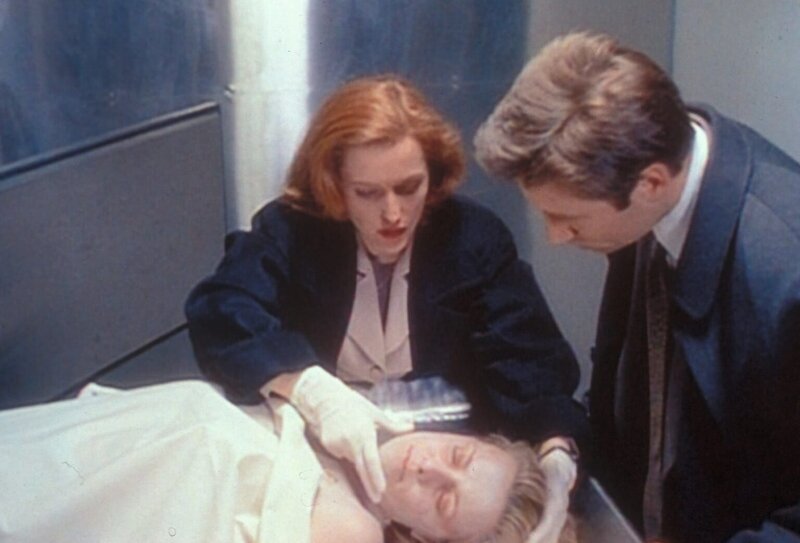 Scully (Gillian Anderson, l.) will Mulder (David Duchovny, r.) eine Phosphoressenz um Mund und Nase einer ermordeten Prostituierten zeigen, die sie bei ihrer ersten Untersuchung entdeckt hat. – Bild: TM + © 2000 Twentieth Century Fox Film Corporation. All Rights Reserved. Lizenzbild frei