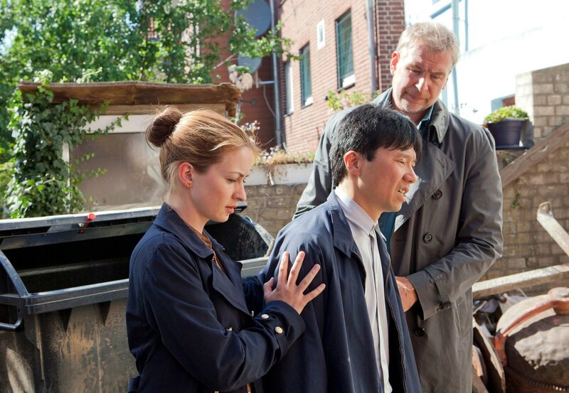 Nora (Loretta Stern, l.) und Jon (Frank Vockroth, r.) stützen Hu (Yuki Iwamoto, M.), den verzweifelten Vater des Ermordeten. – Bild: NDR/​ARD/​Georges Pauly