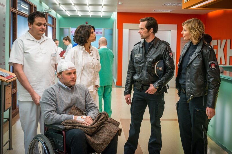 Jasmin (Gerit Kling, m.) klärt Mattes (Matthias Schloo, 2.v.r.) und Melanie (Sanna Englund, r.) bezüglich der Verletzungen ihres Patienten, Nils Steinbach (Mirco Reseg, 2.v.l.), auf. – Bild: ZDF und Boris Laewen