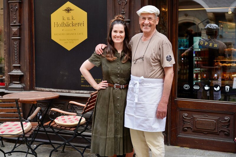 Hoagascht-Moderatorin Conny Bürgler und der ehemalige Bäckermeister und Besitzer Fritz Rath posieren vor der k. und k. Hofbäckerei – Bild: ServusTV / Brigitte Erhart / Degn Film / Apple Photos Clean Up Hoagascht-Moderatorin Conny Bürgler und der ehemalige Bäckermeister und Besitzer Fritz Rath posieren vor der k. und k. Hofbäckerei – Bild: ServusTV / Brigitte Erhart / Degn Film / Apple Photos Clean Up