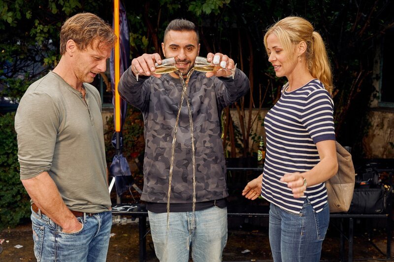 Als Stefan Vollmer (Hendrik Duryn, l.) und Karin Noske (Jessica Ginkel) vor dem Rap Club diskutieren, will Rapper MoTrip das Eis mit einem Prosecco brechen. – Bild: RTL /​ Frank Dicks
